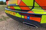 Close-up rear section of a high-visibility police vehicle outdoors, showing orange and neon yellow reflective panels, red POLICE lettering, a blue emergency light, and part of the black bumper and exhaust on a gravel path.